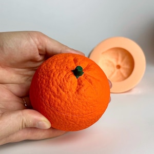 Puede incluir: Una esfera naranja brillante con un tallo verde, que se asemeja a una naranja, sostenida en una mano. Al lado, un molde circular de color naranja claro. La esfera es el foco, con una textura superficial detallada.