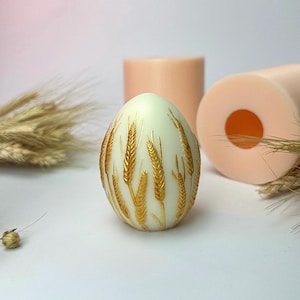 Könnte beinhalten: Eine weiße, eiförmige Kerze mit einem goldenen Weizenmuster. Die Kerze steht auf einer weißen Oberfläche.