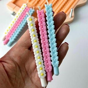 May include: A hand displays several decorative candles. One candle is white with yellow-centred daisies, while others are pink and blue with bow or butterfly designs. The candles are slender and have a string wick.
