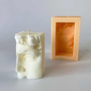 May include: A cream-colored, square candle with a sculpted face design, next to an orange mold of the same face. The candle features a detailed depiction of a classical face, with a wick at the top. The mold is a rectangular shape.