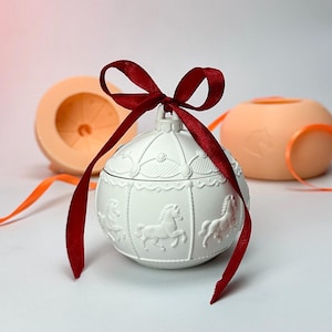 Op de afbeelding: Een witte keramische sieradendoos met carrouselthema en een rode strik. De doos heeft reliëf carrouselpaarden en decoratieve details. Twee perzik-kleurige objecten staan op de achtergrond met oranje linten.