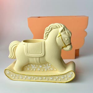 May include: A cream-coloured rocking horse planter with gold accents. The planter features a detailed design with snowflake patterns on the base and gold trim. The horse has a saddle and bridle. An orange mould is in the background.