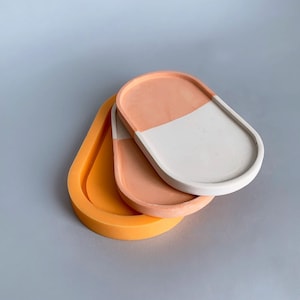 Puede incluir: Tres bandejas decorativas ovaladas en tonos naranja, melocotón y blanco. La bandeja superior está dividida diagonalmente, con melocotón en un lado y blanco en el otro. Las bandejas están apiladas, mostrando sus variados colores y diseños.
