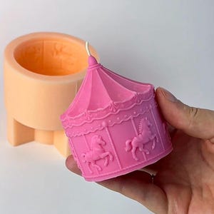 Puede incluir: Una vela rosa con forma de carrusel con una mecha blanca. La vela presenta caballos en relieve y detalles decorativos. Un molde de vela naranja está en el fondo. La vela se sostiene en una mano.