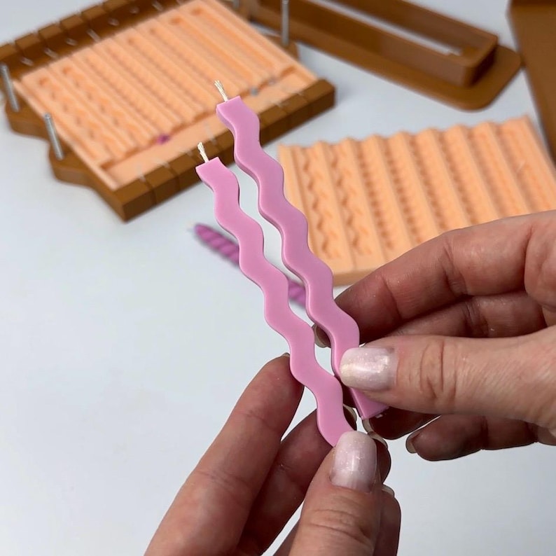 Puede incluir: Dos velas onduladas de color rosa claro con mechas blancas se sostienen. En el fondo, moldes de velas en tonos melocot&oacute;n y marr&oacute;n. Los moldes tienen patrones texturizados para hacer velas. Las velas miden unos 20 cm de largo.