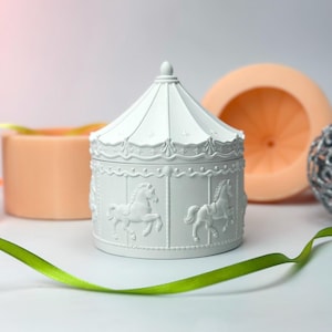 Op de afbeelding: Een witte carrouselvormige sieradendoos met reliëf paardenontwerpen. De doos heeft een conisch dak en is versierd met stervormige accenten. Een groen lint en andere decoratieve items staan op de achtergrond.