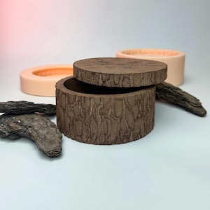 May include: A brown, cylindrical box with a lid, designed to resemble tree bark. The lid is slightly ajar, revealing the interior. Two other containers are in the background, one with a lid and the other without. Decorative wood pieces are scattered around the box.