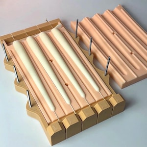 Puede incluir: Un juego de moldes para velas con tres velas blancas en un molde rosa claro. El molde está en un marco marrón claro con varillas metálicas. Otro molde está al lado, con el mismo diseño.