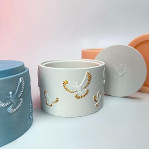 May include: Three decorative containers with lids. The containers are made of white ceramic with a raised design of white doves with gold accents. The lids are white and have a dove design. The containers are in different shades of blue, white, and peach.