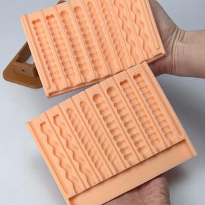 Puede incluir: Dos moldes de silicona color melocot&oacute;n con varios patrones texturizados, que incluyen l&iacute;neas onduladas, c&iacute;rculos y dise&ntilde;os retorcidos. Los moldes son rectangulares y est&aacute;n dise&ntilde;ados para crear elementos decorativos. Un objeto marr&oacute;n est&aacute; en el fondo.