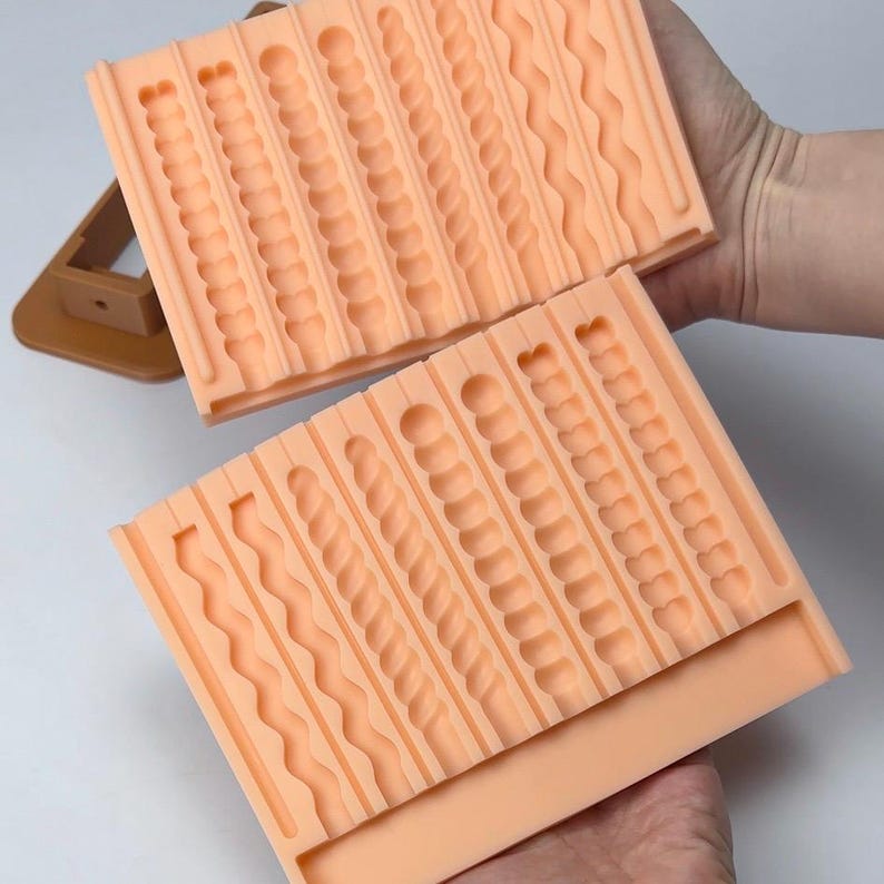 Puede incluir: Dos moldes de silicona color melocot&oacute;n con varios patrones texturizados, que incluyen l&iacute;neas onduladas, c&iacute;rculos y dise&ntilde;os retorcidos. Los moldes son rectangulares y est&aacute;n dise&ntilde;ados para crear elementos decorativos. Un objeto marr&oacute;n est&aacute; en el fondo.