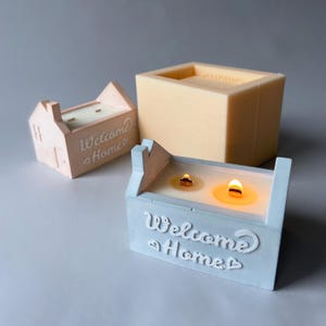 Puede incluir: Tres velas con forma de casa en colores pastel. Una está encendida, mostrando un brillo cálido. Las velas tienen la inscripción "Welcome Home". Un molde rectangular en el fondo.