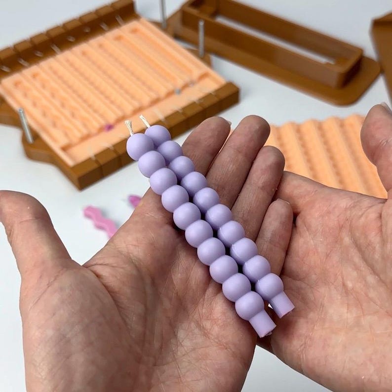 Puede incluir: Dos hebras de velas de cuentas de color lavanda se sostienen en las manos de una persona. Las velas tienen una mecha de cuerda y est&aacute;n junto a moldes de velas. Los moldes son de color marr&oacute;n y melocot&oacute;n.