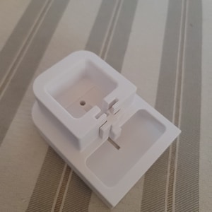 May include: A white plastic device with a rectangular base and a square top section. The top section has a central hole and interlocking tabs. The device is designed to hold something. The background is a striped fabric.