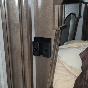 May include: A black plastic phone holder with two star cutouts mounted on a brown wooden wall. The holder is next to a bed with white and brown bedding.