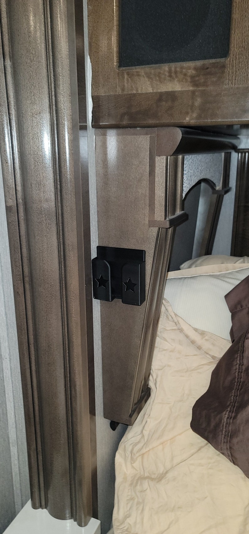 May include: A black plastic phone holder with two star cutouts mounted on a brown wooden wall. The holder is next to a bed with white and brown bedding.