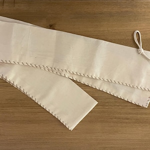 May include: A white fabric roll-up case with brown stitching and a leather strap with a metal buckle. The case is folded in half and the strap is looped around the case.