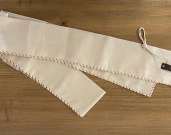 Puede incluir: Un estuche enrollable de tela blanca con costuras marrones y una correa de cuero con una hebilla de metal. El estuche está doblado por la mitad y la correa está enlazada alrededor del estuche.