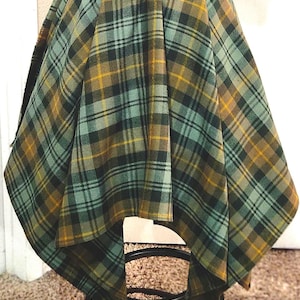 May include: A plaid fabric in shades of green, yellow, and black is draped over a dark brown object. The fabric has a classic plaid pattern. The object appears to be a skirt or a similar garment. The fabric is draped over a black metal stand.