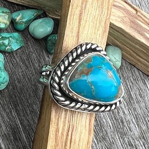 May include: A sterling silver ring with a triangular turquoise stone. The turquoise is a vibrant blue with brown matrix patterns. The ring has a braided rope design around the stone and a polished silver band. Turquoise stones and wooden blocks are in the background.