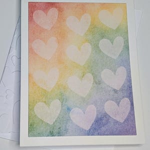 May include: A greeting card featuring a rainbow watercolour background and a grid of white heart shapes. The card is in a white frame, with a matching envelope and a light blue envelope with heart imprints.