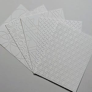 May include: A set of five rectangular white cards arranged in a fan shape. Each card features a distinct embossed pattern, including intersecting circles, concentric rings, geometric triangles, a diagonal diamond grid, and a diamond hatch design.