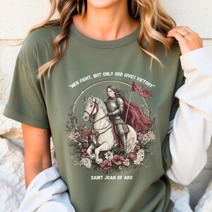 May include: Green t-shirt with a graphic of Saint Joan of Arc riding a white horse. The graphic includes the text "Men fight, but only God gives victory" and "Saint Joan of Arc".