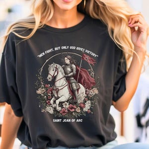 May include: A black t-shirt with a graphic of Saint Joan of Arc riding a white horse. The graphic includes the text "Men fight, but only God gives victory" and "Saint Joan of Arc".