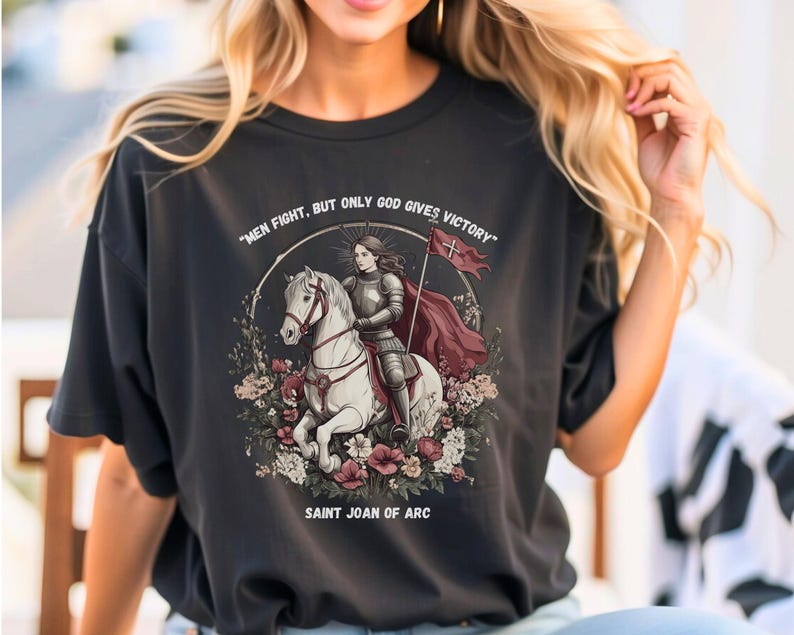 May include: A black t-shirt with a graphic of Saint Joan of Arc riding a white horse. The graphic includes the text "Men fight, but only God gives victory" and "Saint Joan of Arc".