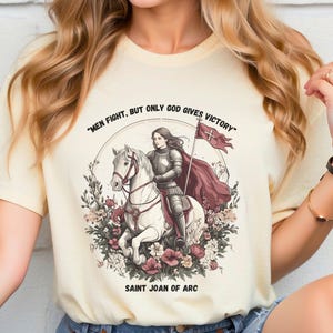 May include: A cream-colored t-shirt with a graphic of Joan of Arc riding a white horse. The graphic includes a red banner with a white cross and the text "Men fight, but only God gives victory." The text "Saint Joan of Arc" is printed below the graphic.