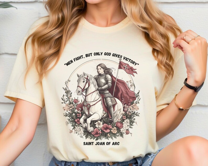 May include: A cream-colored t-shirt with a graphic of Joan of Arc riding a white horse. The graphic includes a red banner with a white cross and the text "Men fight, but only God gives victory." The text "Saint Joan of Arc" is printed below the graphic.