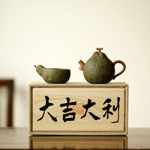 Puede incluir: Una tetera y una taza de té de cerámica verde en forma de pera, sentadas sobre una caja de madera con los caracteres chinos "大吉大利" (que significa "gran fortuna") escritos en tinta negra.