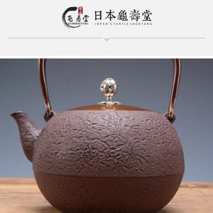 Puede incluir: Una tetera de hierro fundido marrón con una superficie texturizada y una perilla plateada en la tapa. La tetera tiene una manija curva y un pico. El texto "日本龜壽堂 JAPAN'S TURTLE SHOUTANG" está impreso en la parte superior de la imagen.