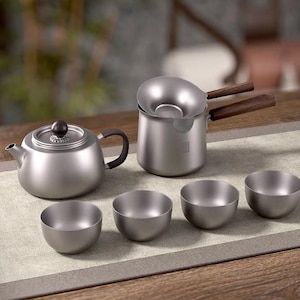 May include: A modern tea set in brushed silver, including a teapot, a strainer pot with wooden handles, and four small bowls. The teapot has a black knob and handle. The set is arranged on a neutral-toned placemat.