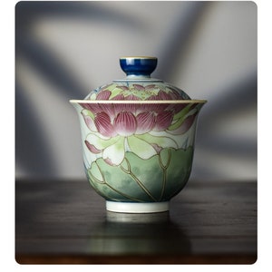 May include: A ceramic tea bowl with a matching lid. The bowl and lid are decorated with a floral design featuring pink lotus flowers and green leaves. The lid has a blue knob. The bowl sits on a white base.