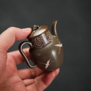 以下が含まれることがあります： 曲線的なハンドルと注ぎ口が付いた、小さな茶色のセラミックティーポット。 蓋には装飾的な雲のデザインがあり、本体には白い鶴のイラストが施されています。 ティーポットは暗い背景に対して手で持たれています。