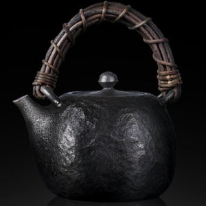 以下が含まれることがあります： 黒色の鋳鉄製の急須で、表面はざらざらしており、持ち手は編み込まれています。急須は丸みを帯びたボディと小さな注ぎ口を持っています。