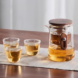 May include: A glass tea set with a wooden lid and handle. The set includes a teapot and three small teacups, all filled with amber tea. The teapot has a built-in infuser. The set is on a light-coloured placemat.