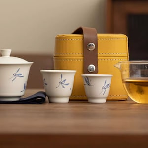 Könnte beinhalten: Eine gelbe Leder-Teedose mit Deckel, zwei weiße Teetassen mit blauen Libellen-Designs und eine klare Glasteekanne mit Henkel, gefüllt mit hellbraunem Tee.