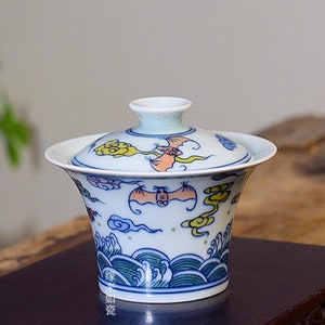May include: A white ceramic teacup with a lid, featuring a blue and green wave pattern and colorful bats and clouds. The teacup is on a dark wooden surface.