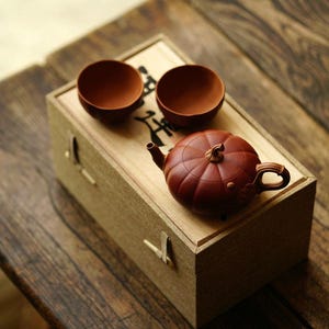 Puede incluir: Una caja de madera marrón con una tetera de cerámica roja con forma de calabaza, dos pequeñas tazas de cerámica roja y caracteres chinos en la caja.