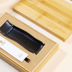 Könnte beinhalten: Ein schwarzes Keramik-Teetablett mit goldenen Rändern, präsentiert in einer Bambusbox. Eine weiße Karte mit goldenem Text ist enthalten. Die Box ist geöffnet und zeigt das Tablett und die Karte. Der Bambusdeckel ist teilweise sichtbar.