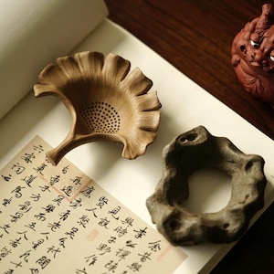 Peut inclure: Deux accessoires à thé en céramique sur un livre avec de la calligraphie chinoise. L'un est une passoire à thé en forme de feuille brune avec un motif de trous. L'autre est un porte-thé en forme de roche grise.