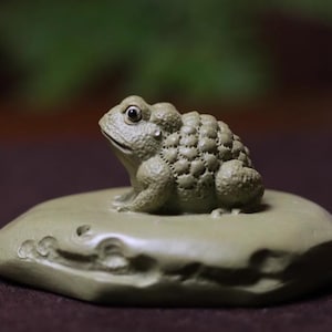 Puede incluir: Una figurita de rana de cerámica verde sentada sobre una roca de cerámica verde. La rana tiene la espalda texturizada y una cara detallada.