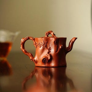 May include: A red clay teapot with a tree trunk design. The teapot has a small spout and a handle that looks like a branch.