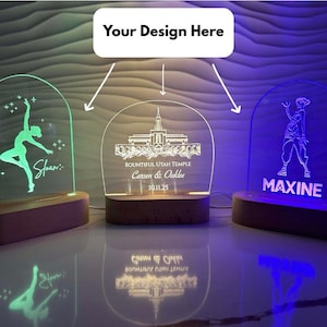 Puede incluir: Tres luces de noche LED personalizadas con bases de madera. Una brilla en verde con una silueta de bailarina y el nombre "Sloan". Otra muestra un templo y nombres. La tercera es azul con una persona y el nombre "MAXINE".