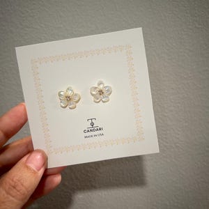 Peut inclure: Boucles d'oreilles en forme de fleur avec une finition nacrée, présentées sur une carte blanche avec une bordure décorative. La carte porte l'inscription "CANDARI MADE IN USA". Chaque boucle d'oreille a un petit centre scintillant.