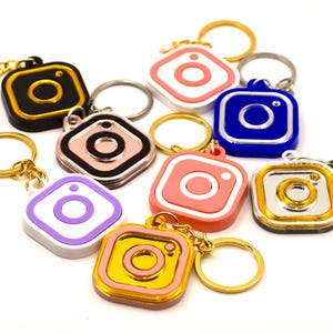 May include: A selection of keyrings in various colours, each featuring a square design with a central camera icon. The keyrings have gold or silver key rings. Colours include black, pink, blue, gold, and purple.