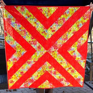 May include: A handmade quilt with a vibrant design. Bold red chevron stripes intersect with floral patterned sections in shades of pink, yellow, and green. The quilt is square-shaped and appears to be machine-stitched.