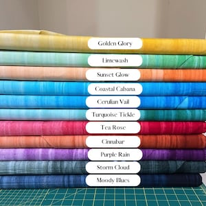 May include: A stack of fabric rolls in various colors, including yellow, green, orange, blue, pink, and purple. Each roll is labeled with a color name such as "Golden Glory", "Limewash", and "Sunset Glow". The fabric is neatly stacked, showcasing a range of hues.